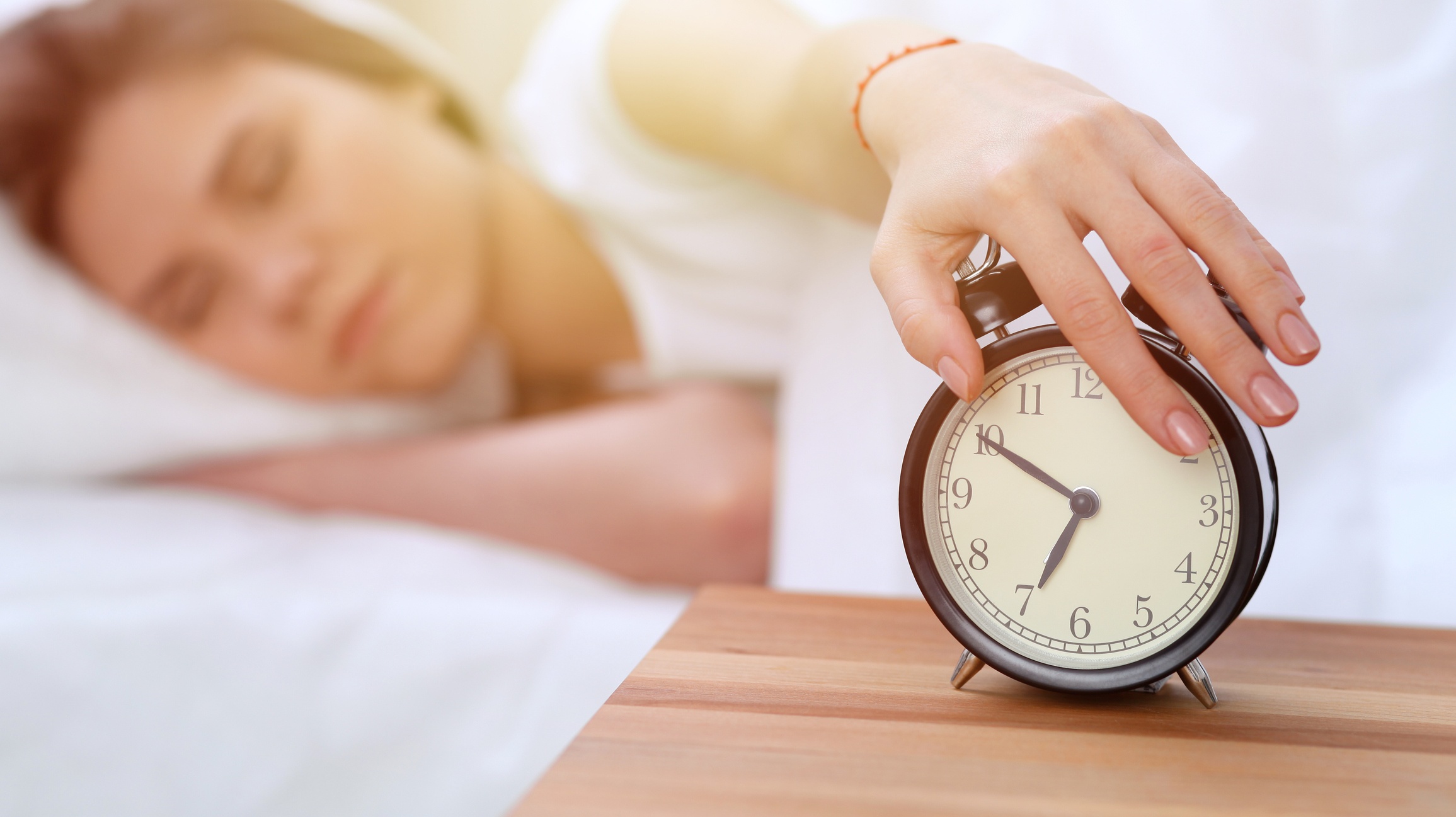 Alarm clock opposite of sleepy young woman stretching hand to ringing alarm willing turn it off. Early wake up, not getting enough sleep concept